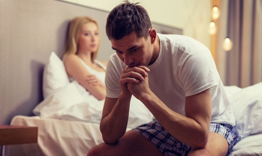 Disfunción erectil y periodontitis podrían estar relacionadas: joven preocupado sentado en la cama con pareja al fondo.